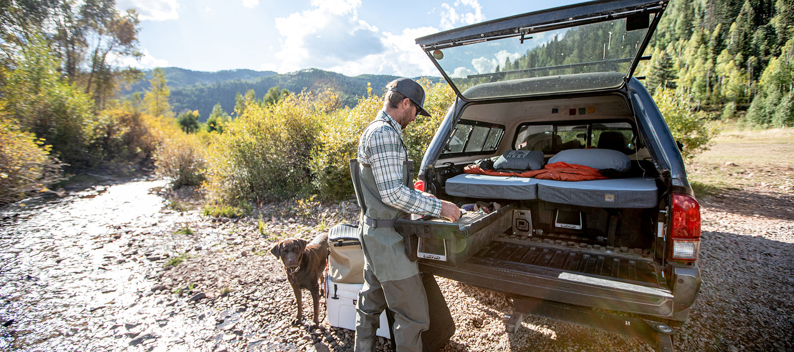 Toyota tacoma next a river, fisherman pulling items out of the back of the truck, dog next to him